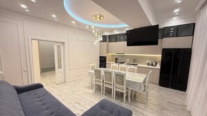 Un salon moderne avec un canapé et un pouf, un coin repas avec une table et des chaises, une cuisine équipée d’appareils électroménagers et un téléviseur à écran plat fixé au mur.
