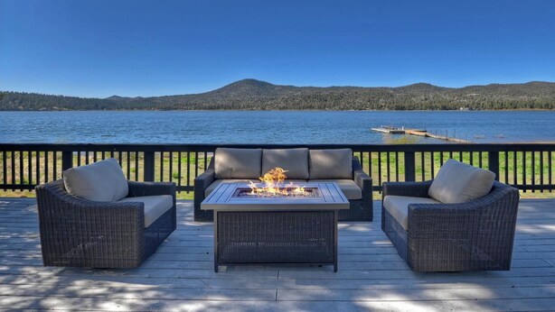 Una terraza con mobiliario de mimbre y una chimenea, con vistas a un lago con muelle y colinas boscosas.