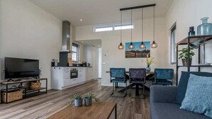 Ein modernes Wohnzimmer mit Sofa, Couchtisch und Fernseher. Es gibt einen Essbereich mit Stühlen und Tisch. Der Raum hat Holzböden und eine Küche im Hintergrund.