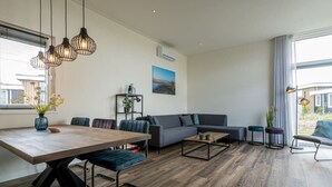 Ein modernes Wohnzimmer mit Esstisch aus Holz, einem grauen Sofa und einem Couchtisch. Es gibt Pendelleuchten, ein Bücherregal und ein Fenster mit Blick.