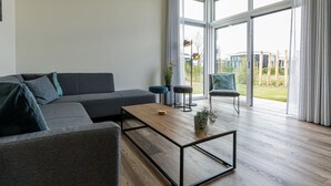 Ein modernes Wohnzimmer mit einem grauen Sofa, einem Holztisch und Blick auf einen Garten durch große Fenster.