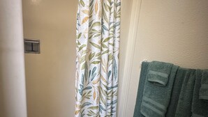 Ein Badezimmer mit Duschvorhang mit Blattmuster, einem Handtuchhalter mit gefalteten Handtüchern und einem Lichtschalter an der Wand.