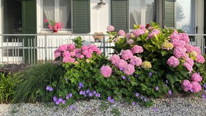 Una casa con balcón, contraventanas verdes y un jardín con flores rosas y púrpuras.