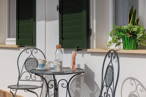 Una pequeña mesa al aire libre con dos sillas, una planta en maceta y una ventana con contraventanas verdes.