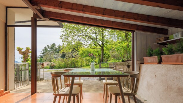 Un patio al aire libre con una mesa de vidrio y sillas de madera, rodeado de un jardín con árboles y plantas.