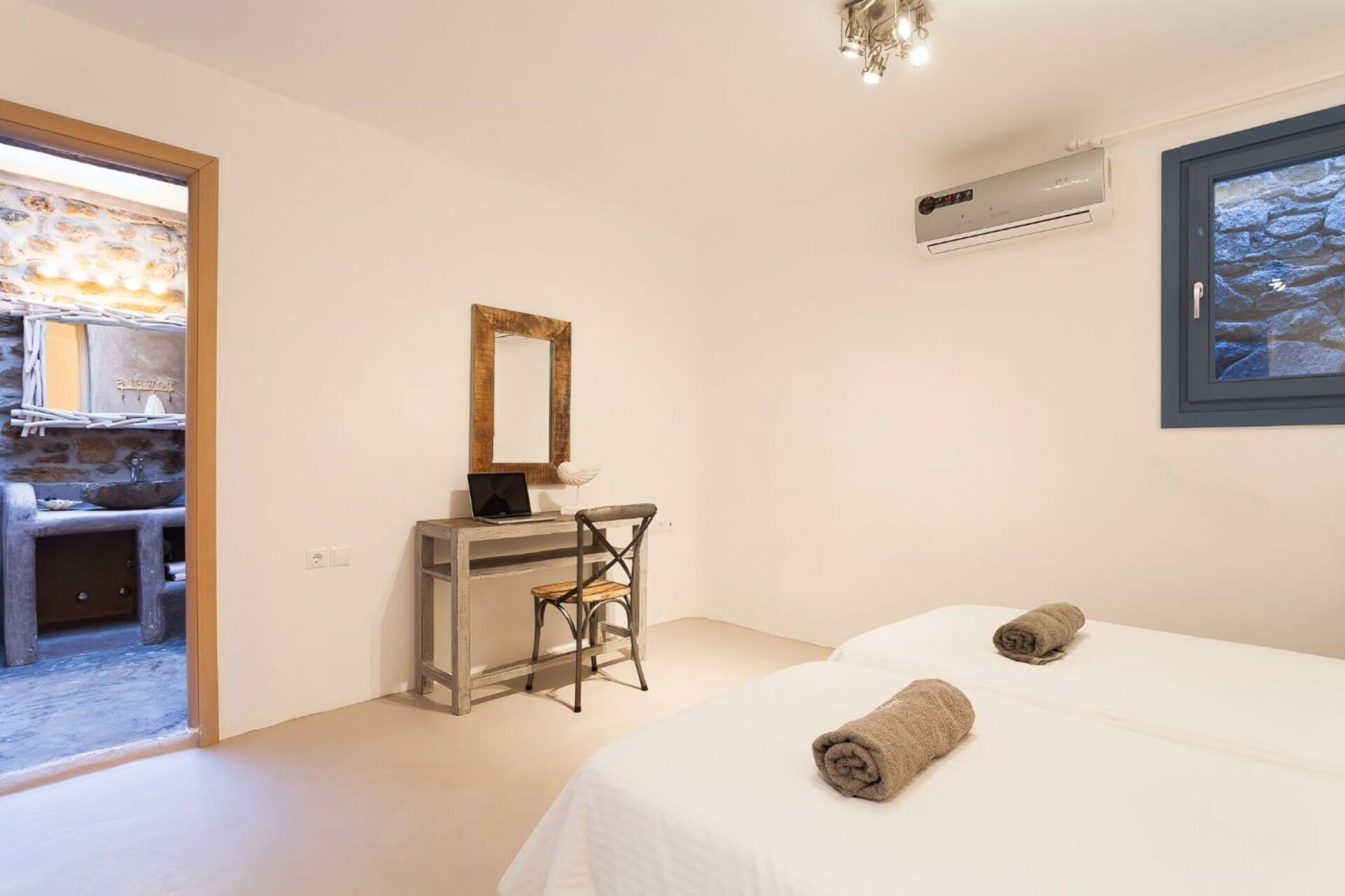 Un dormitorio con cama, un escritorio con silla, un espejo, un equipo de aire acondicionado y una ventana con vista a un muro de piedra.