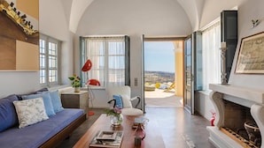 Ein geräumiges Wohnzimmer mit Sofa, Couchtisch und Kamin. Durch eine geöffnete Tür ist ein Balkon mit Blick auf die Landschaft zu sehen.