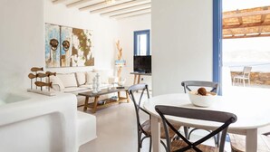 A modern living room with a white sofa, a dining table with chairs, a TV, and artwork on the walls. - My Rental Homes - Villa Paradeisos with private pool and sea view (Mykonos)