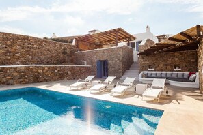A pool area with lounge chairs, a stone wall, and a white building with a wooden pergola.