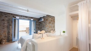 A bedroom with a stone wall, a bed with white linens, a small table with decorative items, and a view of the outdoors. - My Rental Homes - Villa Paradeisos with private pool and sea view (Mykonos)