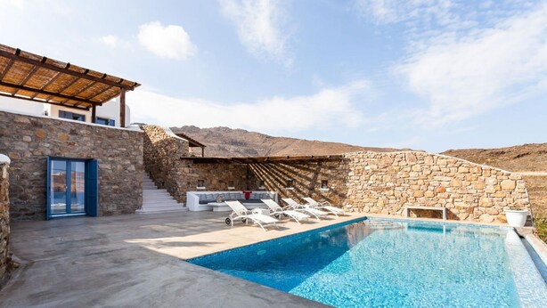 A pool area with stone walls, a covered seating area, and a staircase leading to a second level. - My Rental Homes - Villa Paradeisos with private pool and sea view (Mykonos)
