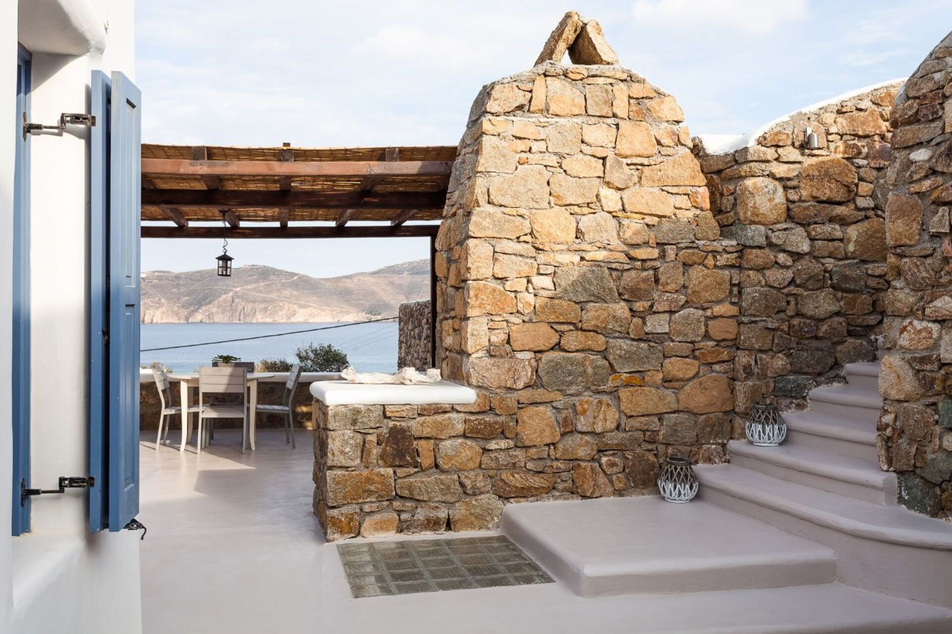 Un espace extérieur entouré de murs en pierre, avec une table et des chaises, un banc en pierre, et une vue sur la mer et les montagnes.