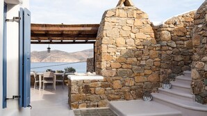 A stone-walled outdoor area with a table and chairs, a stone bench, and a view of the sea and mountains. - My Rental Homes - Villa Paradeisos with private pool and sea view (Mykonos)