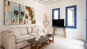 A living room with a white sofa, a coffee table, a TV on a stand, and a large abstract painting on the wall. - My Rental Homes - Villa Paradeisos with private pool and sea view (Mykonos)