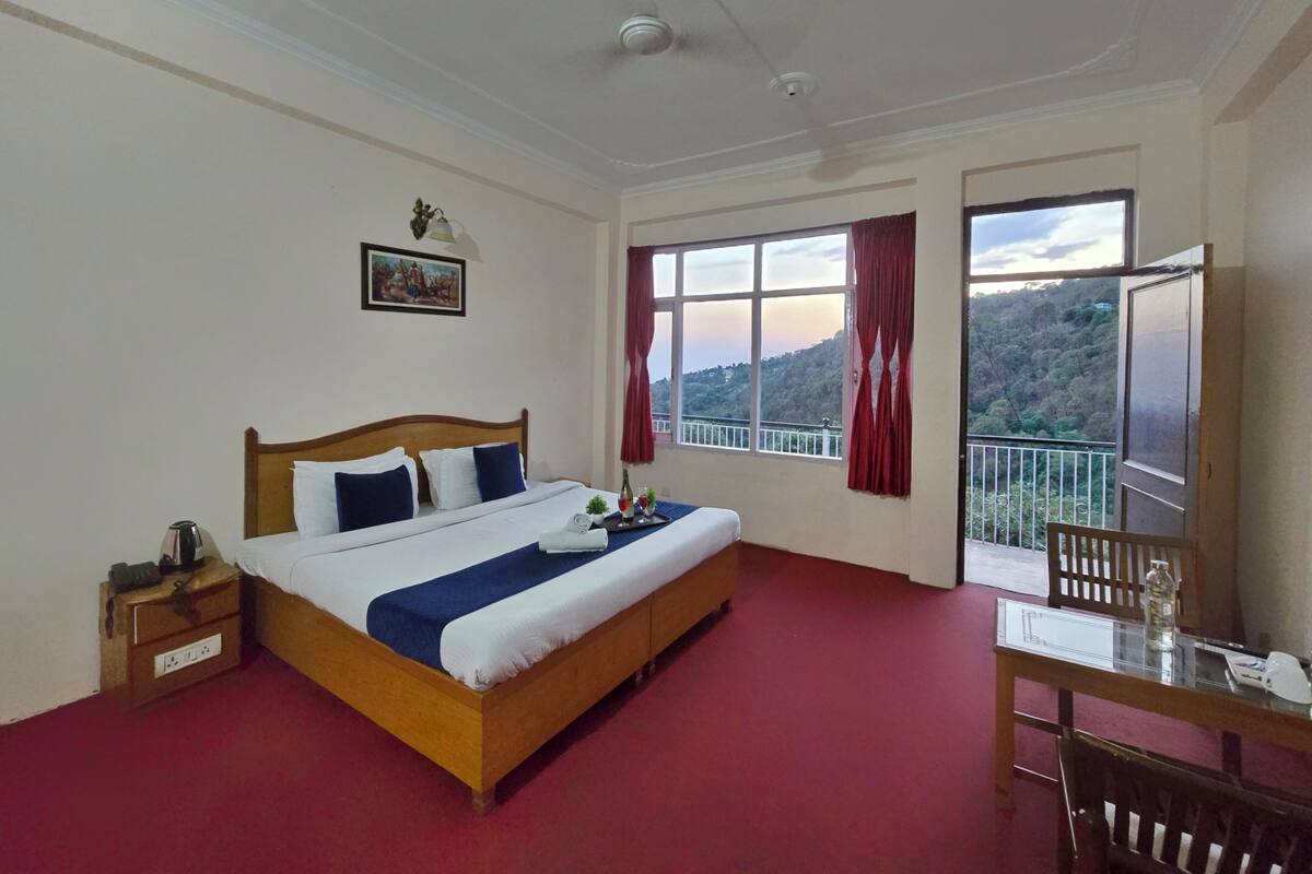 A hotel room with a large bed, a desk with a chair, a window with a view of the hills, and a ceiling fan.