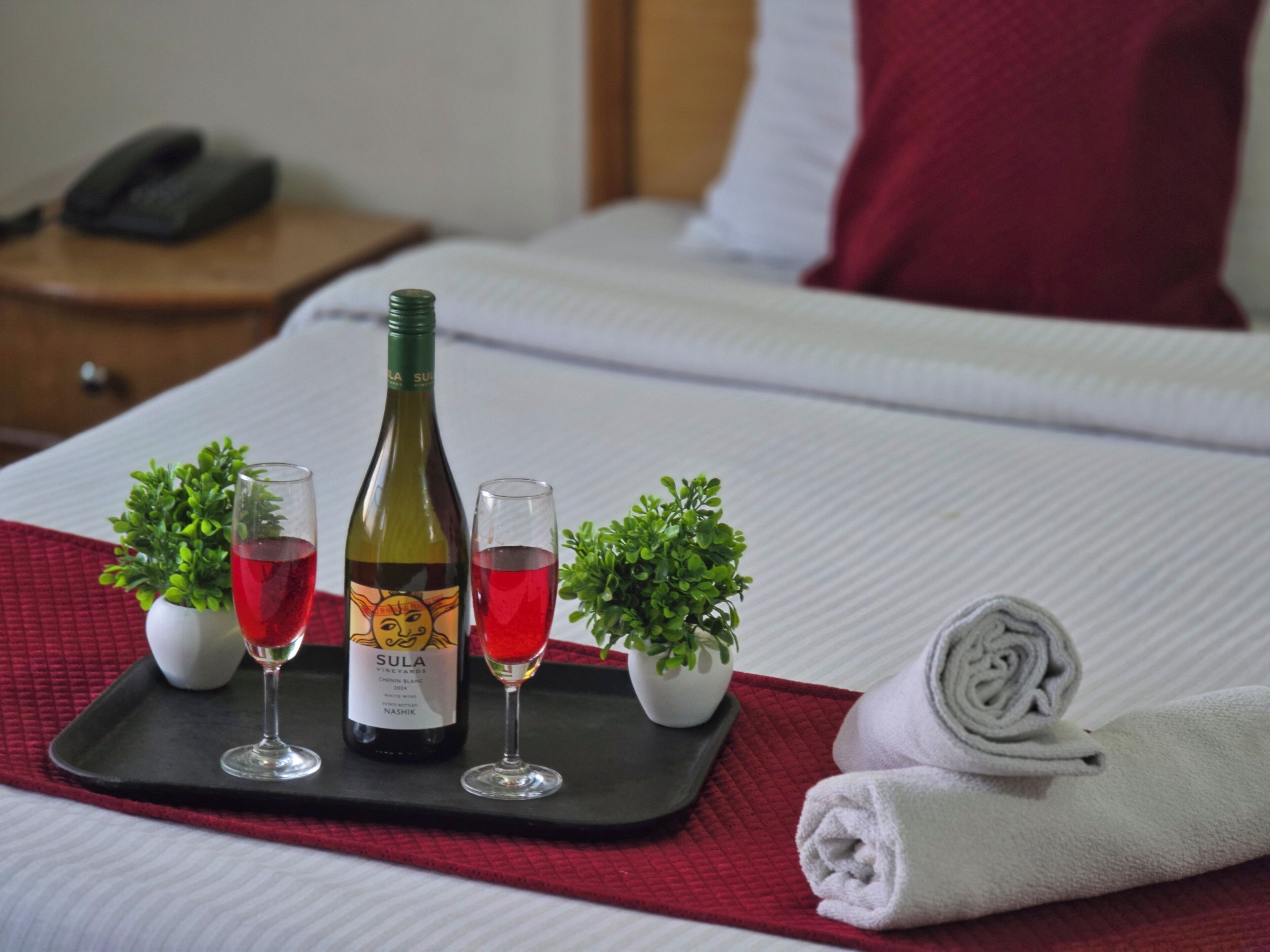 Un lit bien fait, recouvert d’une couverture rouge, une bouteille de vin Sula Vineyards, deux verres de vin rouge et de petites plantes en pot disposées sur un plateau.