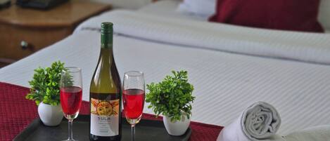 Un lit bien fait, recouvert d’une couverture rouge, une bouteille de vin Sula Vineyards, deux verres de vin rouge et de petites plantes en pot disposées sur un plateau.