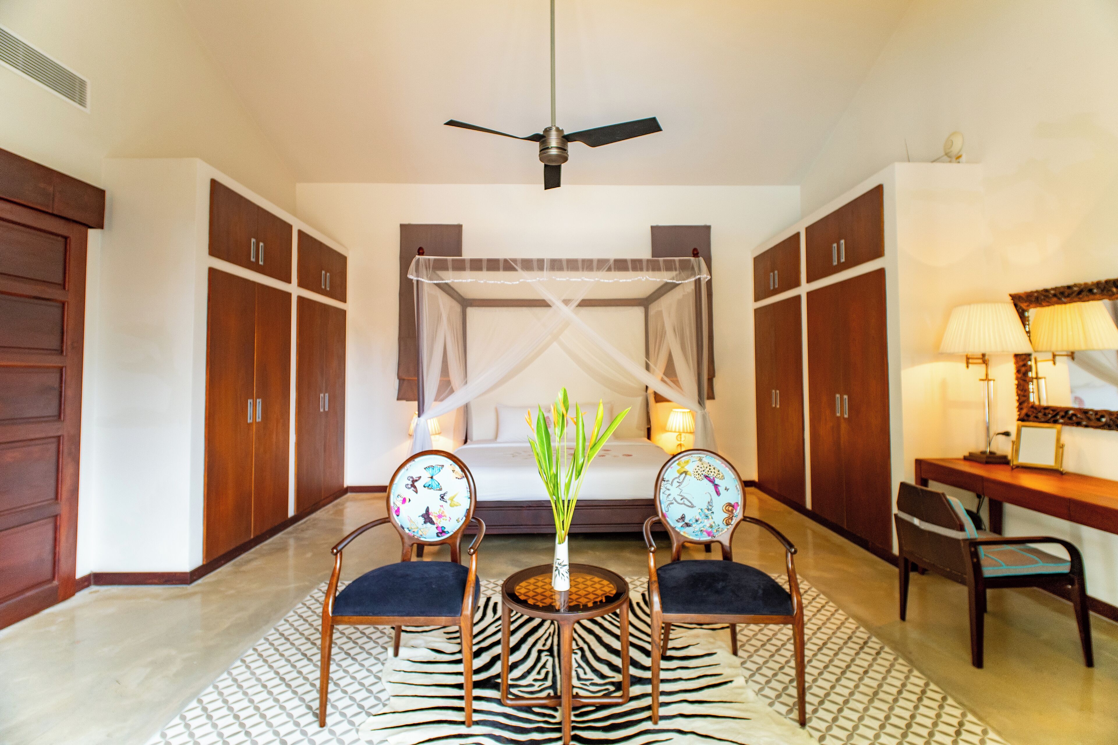 A bedroom with a four-poster bed, a ceiling fan, two chairs with butterfly designs, a small round table with a vase, and a desk with a lamp.