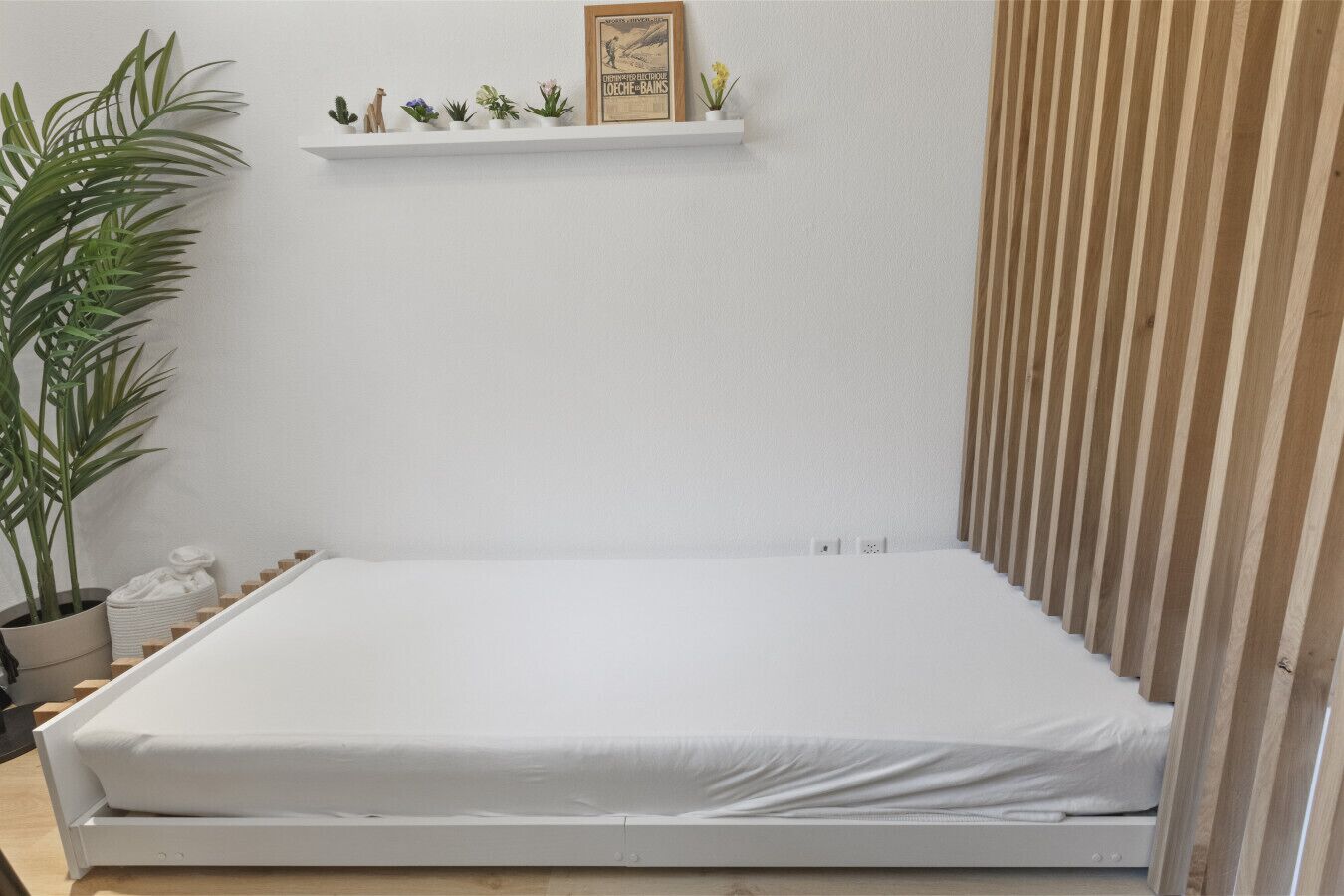 Ein schlicht eingerichtetes Schlafzimmer mit einem weißen Bett, einer Ablage mit kleinen Pflanzen und einer Wand aus Holzleisten.