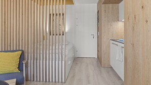 A modern, minimalist room with a wooden slatted partition, a bed, a small table, and a kitchen area with a counter and hanging towel. - Élégant studio, proche des bains & nature (Leukerbad)