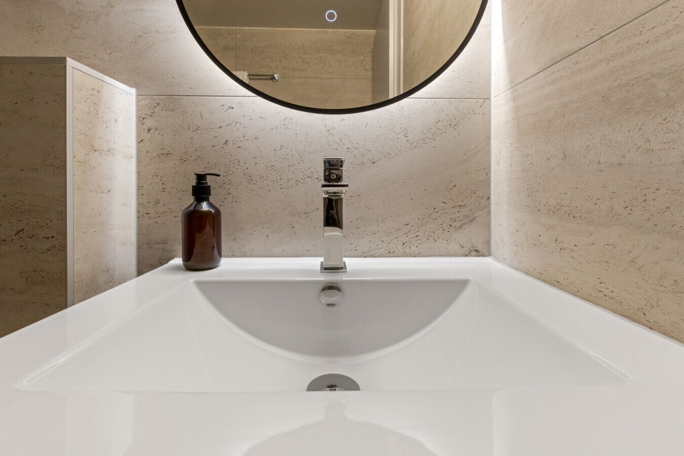 Een moderne badkamer met een wit wastafeltje, een ronde spiegel en een zeepdispenser.