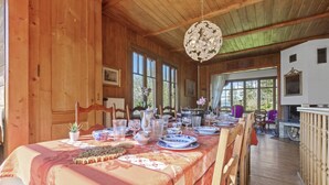 A dining area with a long table set for a meal, wooden walls, large windows, and a hanging light fixture.