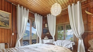 A wooden cabin bedroom with a large bed, two windows with curtains, a wooden ceiling, and a view of mountains.