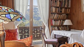 A cozy room with a wooden bookshelf, a table with a lace tablecloth, and a view of a mountainous landscape through a large window.