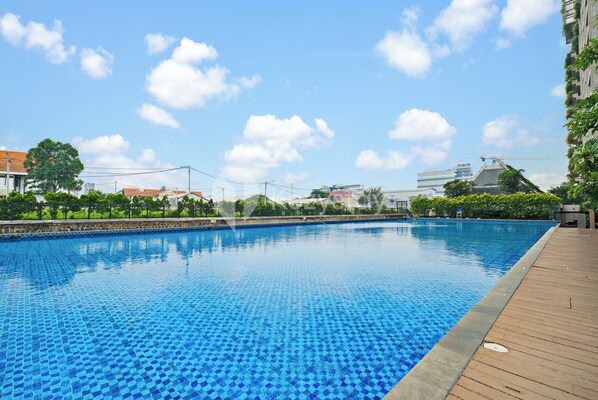 A swimming pool with a wooden deck, surrounded by greenery and buildings in the background. - Kiara by Kozystay - Paskal (Bandung)