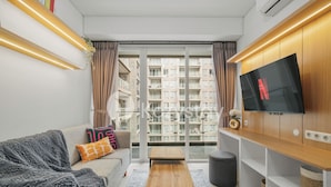 A modern living room with a sofa, a coffee table, a TV on a wooden cabinet, and a view of a residential building through large windows. - Kiara by Kozystay - Paskal (Bandung)