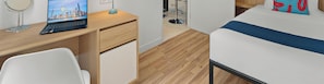 Habitación pequeña con escritorio de madera, una laptop, una silla, cama con franja azul y una vista urbana en la pantalla de la laptop.