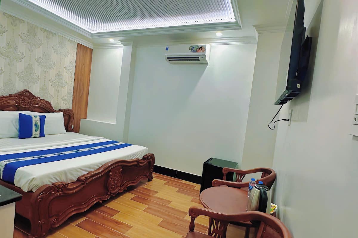 A hotel room with a bed, a small table, a chair, a TV, and an air conditioning unit.