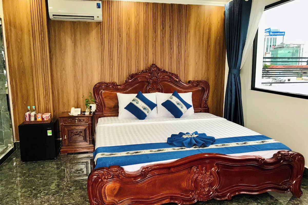 A hotel room with a wooden paneled wall, a large bed with a wooden headboard, a minibar, and a view of a building outside.