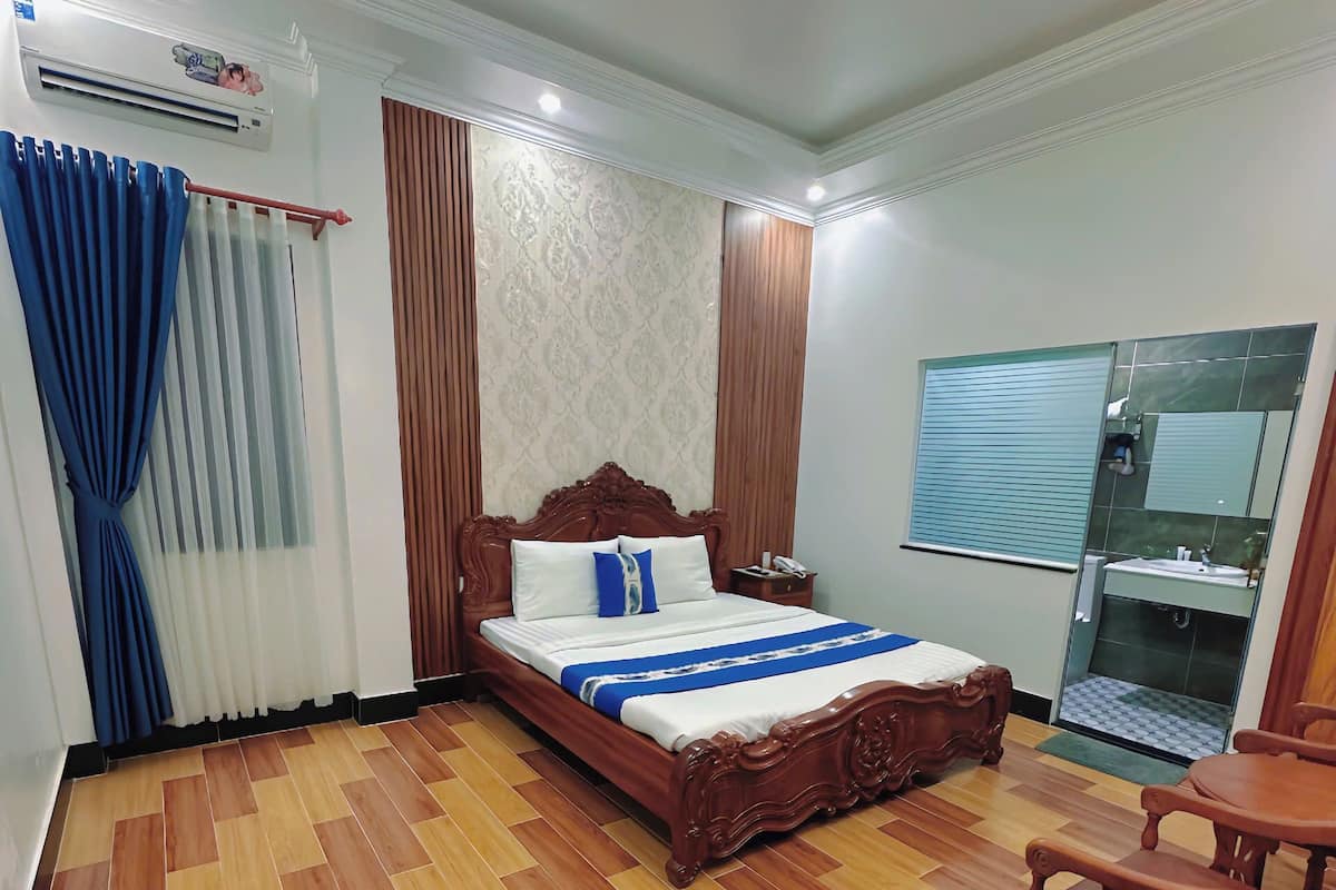 A hotel room with a wooden floor, a bed with a blue and white bedspread, a wooden headboard, a bedside table with a lamp, a window with blinds, and a small table with a chair.