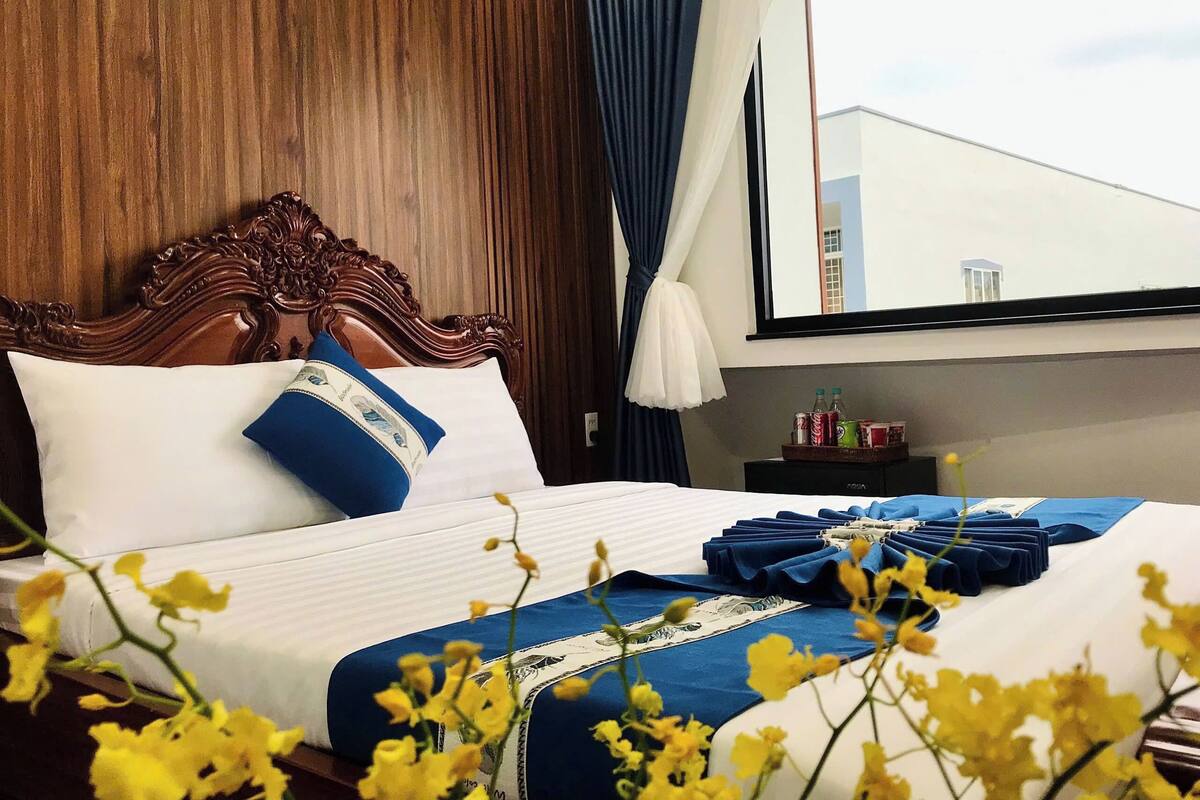 A hotel room with a wooden headboard, white and blue bedding, and a view of the sky through the window.