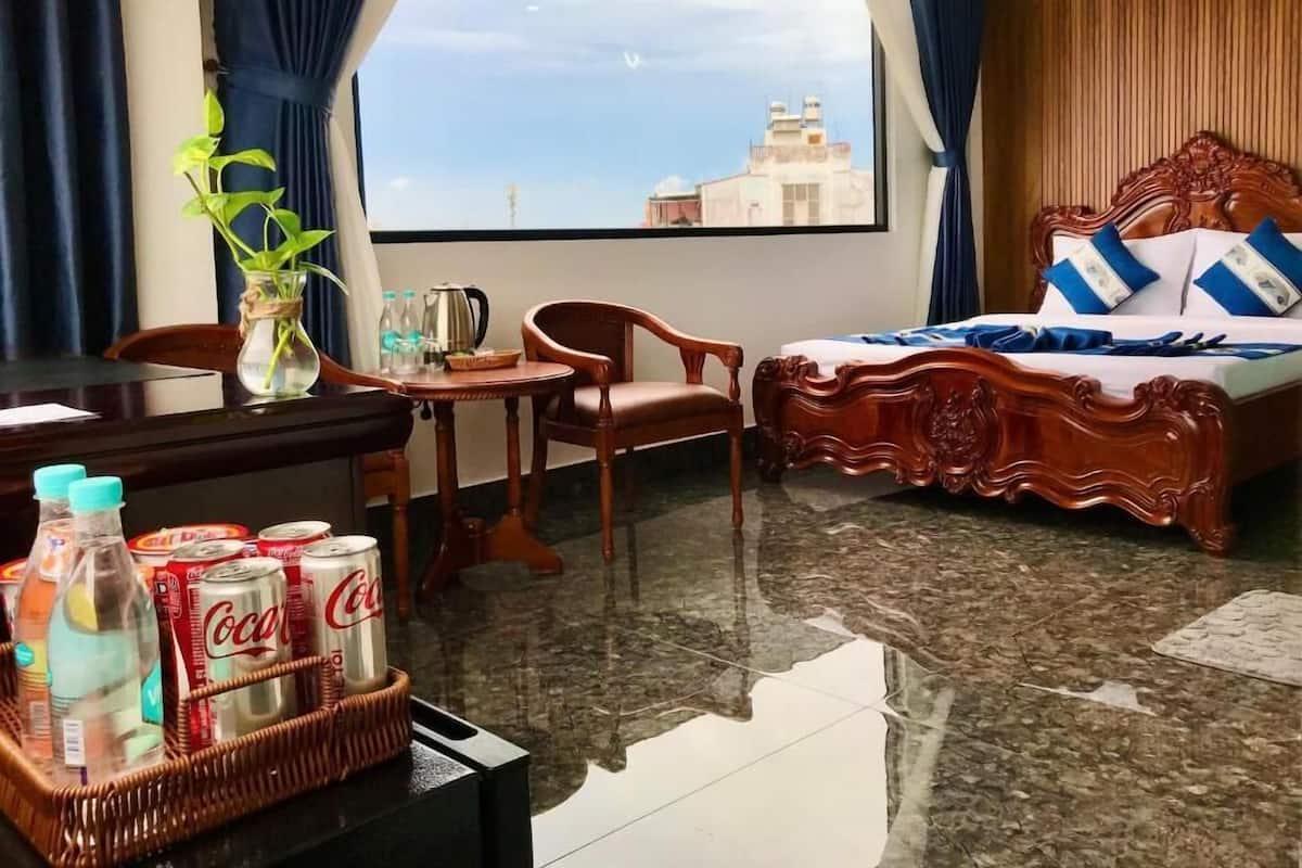 A hotel room with a bed, a desk with a chair, a small table, a potted plant, and a view of buildings through a large window.