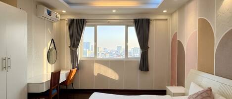 Quarto de hotel com cama, escrivaninha, cadeira, janela com cortinas, ar condicionado e vista para a paisagem urbana.