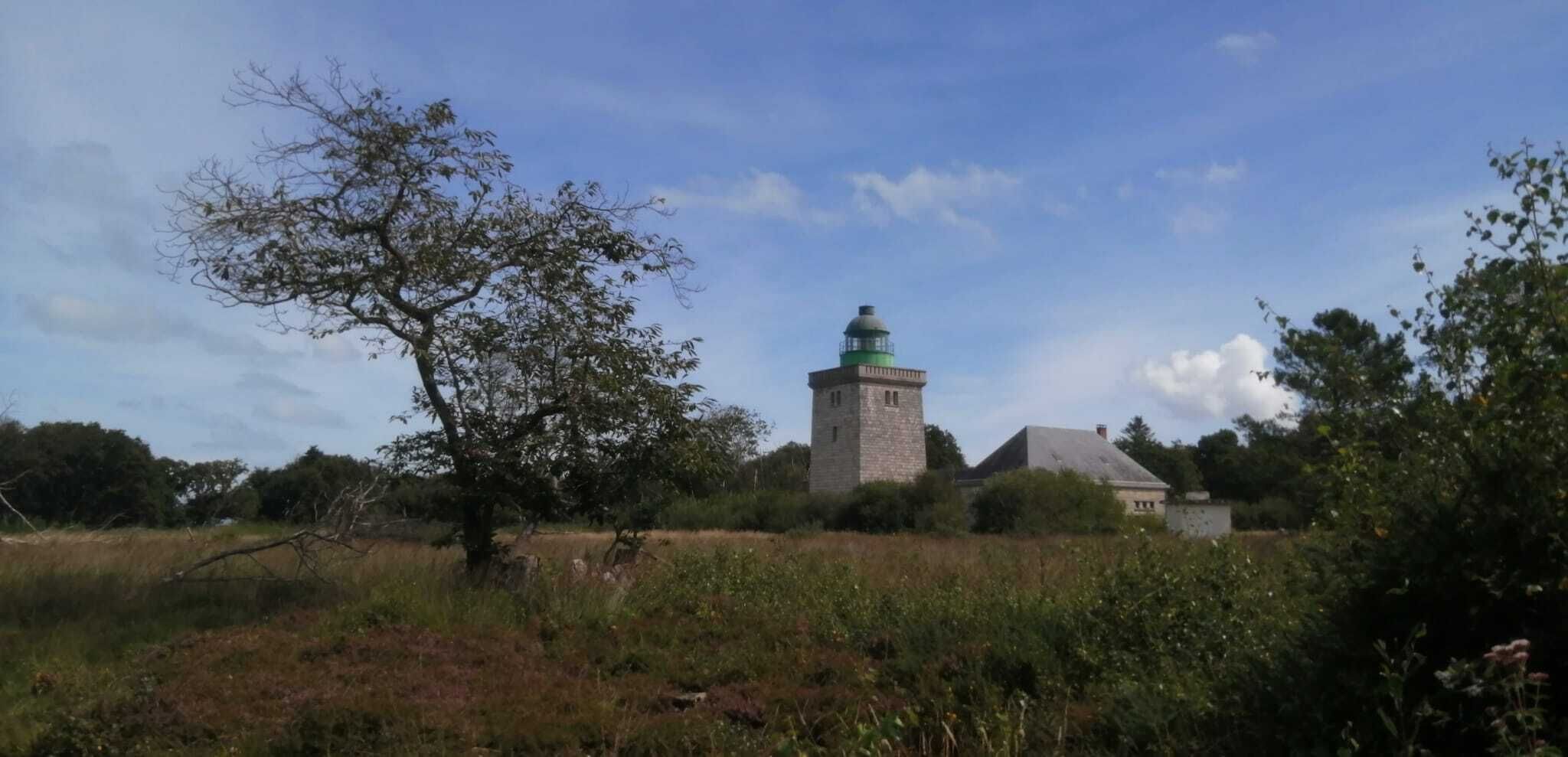Un faro con base de piedra y cúpula verde, rodeado por un campo cubierto de hierba y árboles.