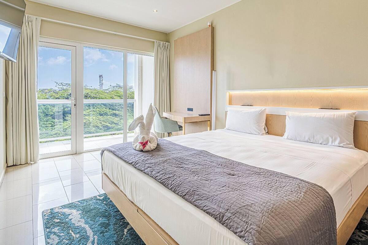 A hotel room with a large bed, a nightstand, a lamp, and a view of greenery through the window.