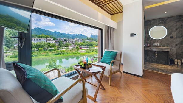 A modern hotel room with a large window offering a view of a lake and mountains, a cozy seating area with a table, and a bathroom with a marble wall and a round mirror. - Riverside Peak Lodge Zhangjiajie Wulingyuan National Park (Zhangjiajie)