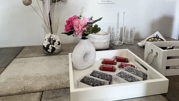 A room with a white wall, a wooden floor, a white table with a vase of flowers, a white tray with chocolates, and a wall with framed pictures.