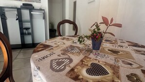 A dining area with a table covered in a patterned tablecloth, chairs, a potted plant, and a wall-mounted clock.
