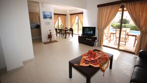 A living room with a TV, a dining area, and a view of a pool. - Seaside 2-bedroom Villa With Private Pool, Garden & Wifi Your Perfect Escape! (Pissouri)
