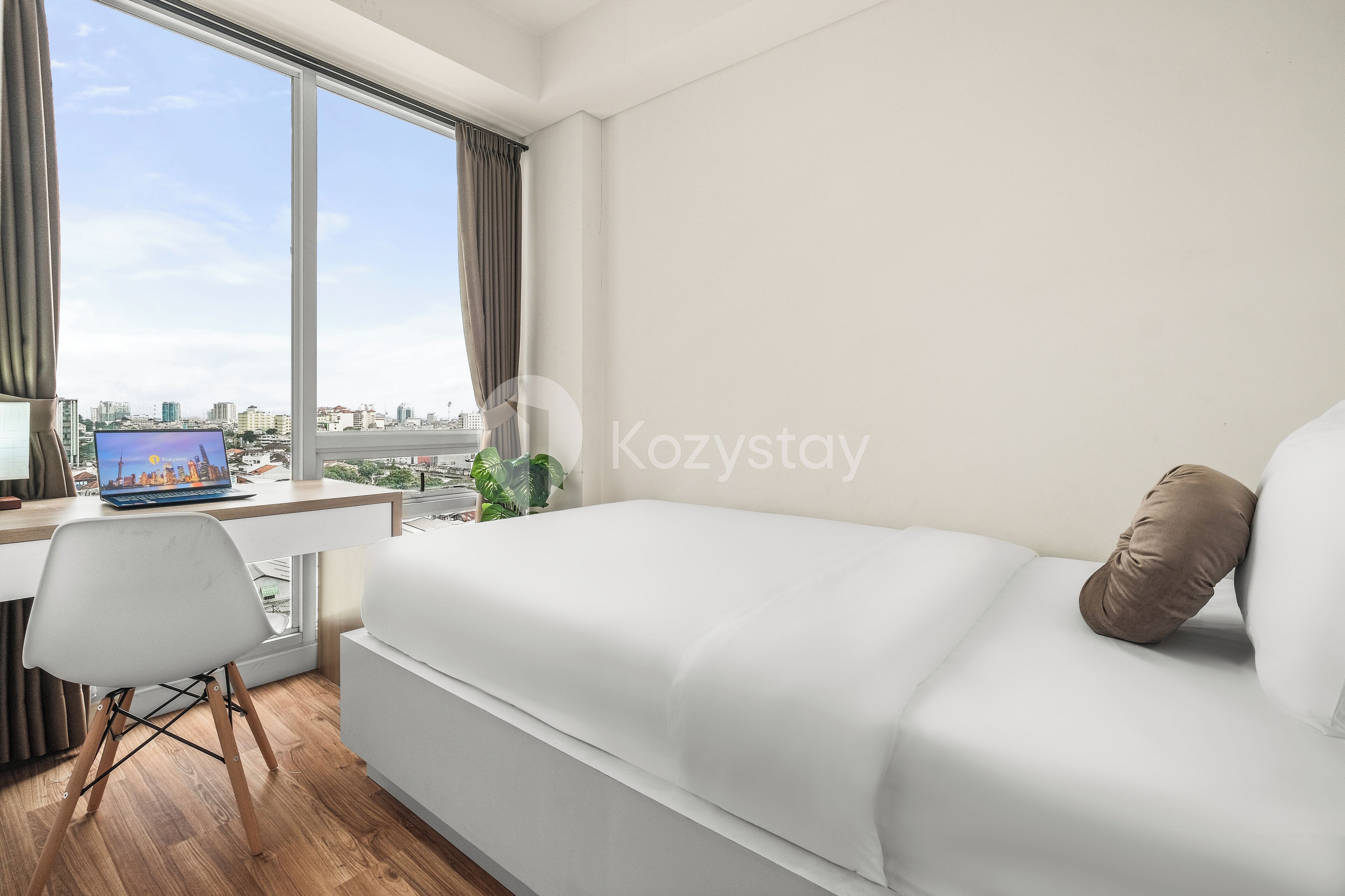 A modern hotel room with a large bed, a desk with a laptop, and a view of the cityscape through large windows.