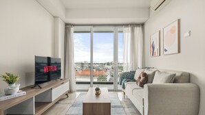 Una sala de estar moderna con un televisor mostrando 'NETFLIX', un sofá, una mesa de centro y vistas a la ciudad a través de amplias ventanas.