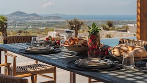 A table set for breakfast with bread, fruit, and a glass of water, overlooking a scenic landscape.