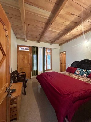 A bedroom with a wooden ceiling, a bed with a red bedspread and floral pillows, a wooden desk with chairs, and a window with curtains. - New Brahma Guesthouse (Sosan)