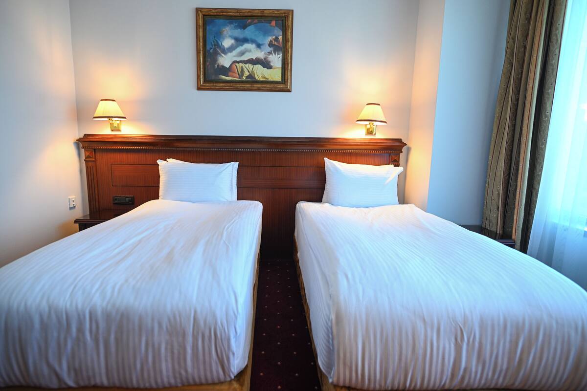 A hotel room with two single beds, a wooden headboard, two wall-mounted lamps, and a painting above the headboard.