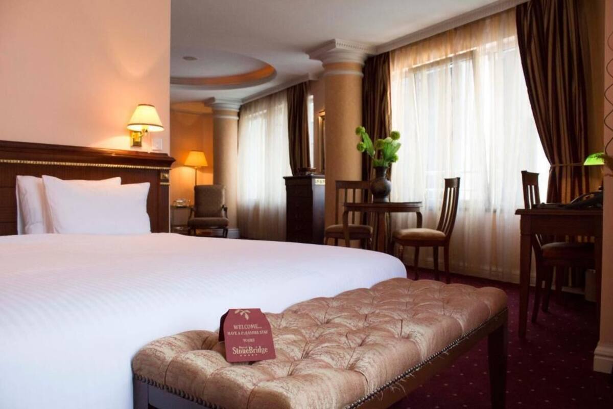 A hotel room with a large bed, a bench, a desk, and a chair. There is a sign on the bench that reads "Welcome StoneBridge."