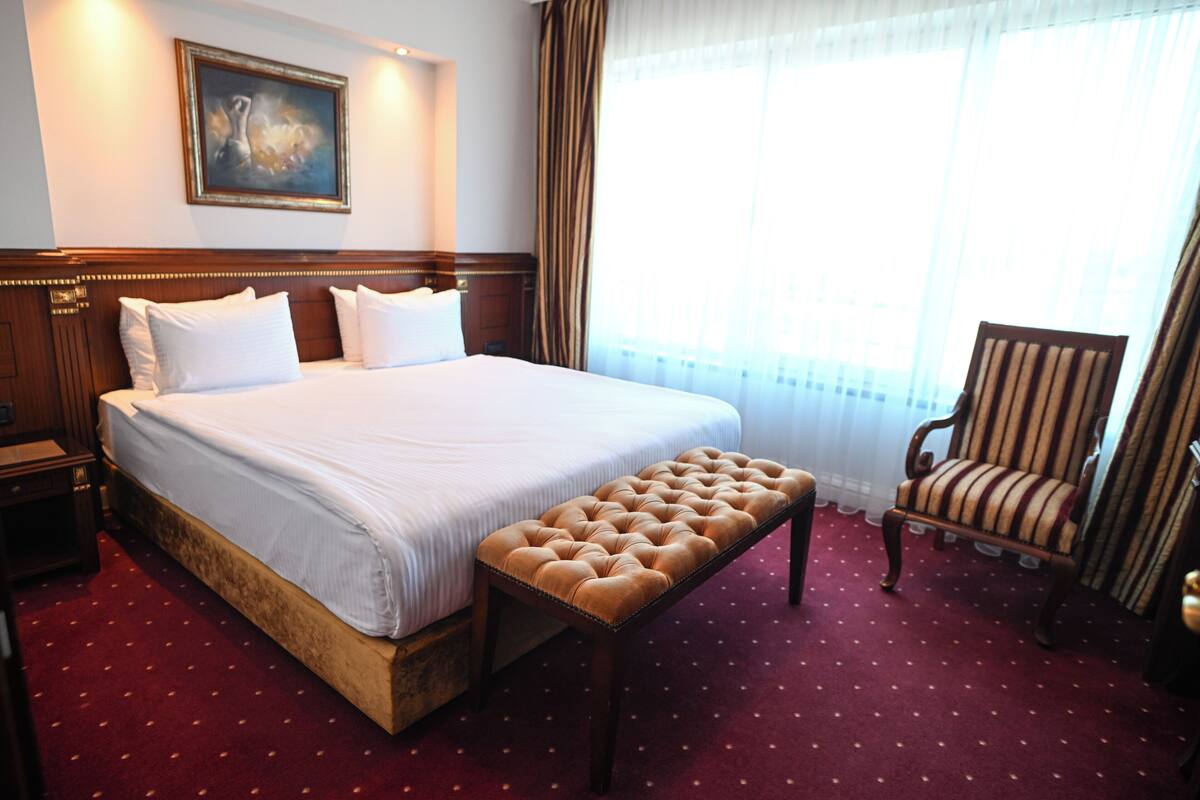 A hotel room with a large bed, a wooden bench, a striped armchair, and a painting on the wall.